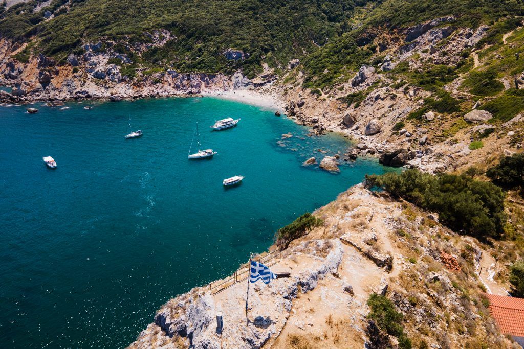 Kastro Beach Skiathos 2026 – historic cliffside beach with turquoise waters and Greek flag view