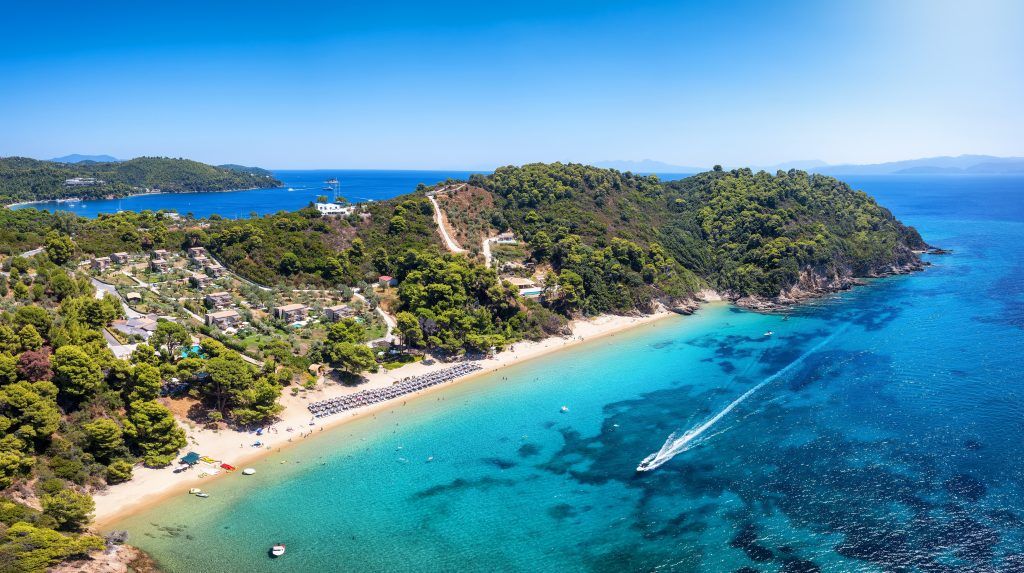 Agia Eleni Beach Skiathos with golden sand, calm waters, and pine trees at sunset.